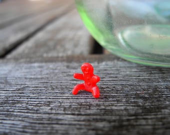 Może przedstawiać: Mała, jaskrawoczerwona plastikowa figurka przedstawiająca postać w pozycji siedzącej z nogami wyciągniętymi do przodu i ramionami rozłożonymi na boki. Figurka ma gładką, formowaną powierzchnię oraz prostą, zaokrągloną głowę z subtelnymi detalami.