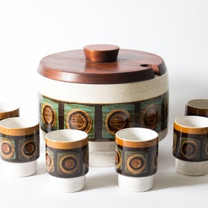 Puede incluir: Un juego de servir de cerámica vintage con un recipiente grande con tapa y seis tazas a juego. El juego presenta una tapa de madera marrón y una base color crema con un diseño geométrico en tonos marrones, turquesa y dorados. Las tazas tienen un diseño similar.