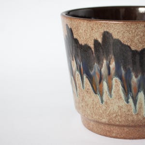 Può includere: Un vaso di ceramica con esterno beige maculato e interno marrone scuro. Il vaso presenta un motivo decorativo di striature verticali blu scuro, nere e arancioni. La base è di colore marrone chiaro uniforme.