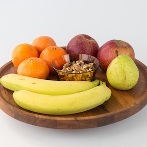 Puede incluir: Una bandeja de madera con frutas frescas y nueces. La bandeja contiene naranjas, manzanas rojas, una pera verde y dos plátanos. Un pequeño cuenco de cristal contiene nueces. La disposición se encuentra sobre un fondo blanco.