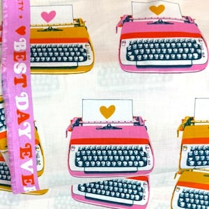 Könnte beinhalten: Weißer Stoff mit einem sich wiederholenden Muster aus rosa und gelben Schreibmaschinen, jede mit einem herzförmigen Papier. Ein rosa Band mit dem Text "BEST DAY EVER" und "RUBY STAR SOCIETY" befindet sich im Vordergrund. Der Stoff ist aus 100% Baumwolle und in Japan hergestellt.