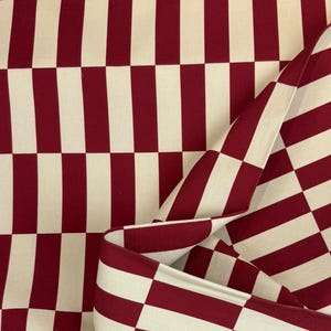 Könnte beinhalten: Nahaufnahme eines Stoffes mit einem auffälligen Schachbrettmuster. Das Design zeigt abwechselnd vertikale und horizontale Streifen in einem tiefen Burgunderrot und Creme. Der Stoff ist gefaltet und zeigt die Kontinuität des Musters.