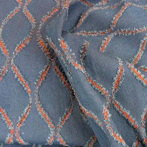 Könnte beinhalten: Nahaufnahme eines blauen Stoffes mit einem orangefarbenen Rautenmuster. Der Stoff hat eine strukturierte Optik mit ausgefransten Kanten entlang der orangefarbenen Nähte, was einen einzigartigen visuellen Effekt erzeugt. Der Stoff ist gefaltet und zeigt Tiefe und Dimension.