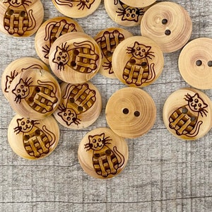 Könnte beinhalten: Eine Sammlung von 14 Holz-Knöpfen mit Katzenmotiv. Die Knöpfe sind hellbraun und haben jeweils zwei Löcher. Die Knöpfe sind in einem Kreis auf einer hellbraunen Holzoberfläche angeordnet.