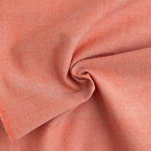 Könnte beinhalten: Nahaufnahme eines gefalteten orangefarbenen Stoffstücks. Der Stoff hat ein strukturiertes Aussehen mit einer leichten Farbvariation, die eine subtile visuelle Tiefe erzeugt. Die Kanten des Stoffes sind sichtbar und zeigen den rohen, unfertigen Rand.