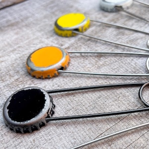 Könnte beinhalten: Fünf Flaschenverschlüsse, jeder in einer anderen Farbe lackiert, sind an Sicherheitsnadeln befestigt. Die Farben sind weiß, gelb, orange, schwarz und grau.