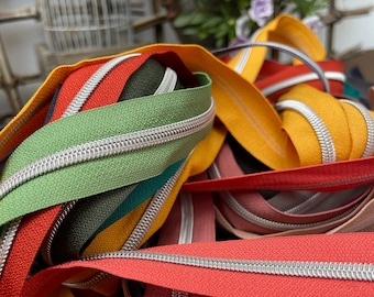 May include: A close-up of a pile of colorful zipper tapes with silver teeth. The tapes are arranged in a random pattern, with some of the tapes overlapping each other. The colors of the tapes include red, orange, yellow, green, blue, and purple.