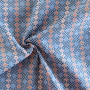 Könnte beinhalten: Nahaufnahme eines blauen Stoffes mit einem geometrischen Muster aus orangefarbenen und grauen Quadraten. Der Stoff ist gefaltet und zeigt seine Textur und sein Design. Das Wort "FABLEISM" ist in Weiß unten rechts aufgedruckt.