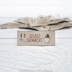 Könnte beinhalten: Beige Stoffetikett mit gestickten Gartengeräten, dem Text "SELBST GEMACHT" und einer Topfpflanze. Weitere Etiketten sind im Hintergrund unscharf. Das Etikett liegt auf einer weißen Holzoberfläche.