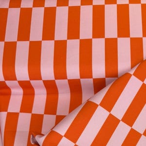 Könnte beinhalten: Gefalteter Stoff mit einem sich wiederholenden Muster aus orangefarbenen und weißen Karos. Das Design des Stoffes zeigt ein Gitter aus orangefarbenen und weißen Quadraten und Rechtecken. Geeignet für Kleidung oder Heimdekoration.