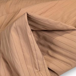 Könnte beinhalten: Nahaufnahme eines gefalteten Stoffes in hellem Braunton mit einem plissierten Design. Vertikale Linien verlaufen über das Material und erzeugen Textur und Schatten. Die Falten betonen den Fall und die Struktur des Stoffes.