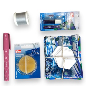 Könnte beinhalten: Ein Set Nähzubehör, bestehend aus einer Spule grauen Fadens, einem rosa Stoffmarker, einem kleinen runden Behälter mit Nähwachs, einer Packung Nadeln und einem Stapel Stoffquadraten mit einem blauen und weißen Blumenmuster.
