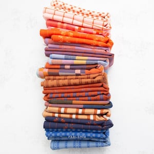 Könnte beinhalten: Ein vertikaler Stapel gefalteter Stoffstücke mit einer Vielzahl von Webmustern und Farben, darunter Pfirsich, Orange, Rost, Violett, Blau und Marineblau. Das Wort FABLEISM ist in kleinen weißen Buchstaben auf dem unteren blauen Stoff aufgedruckt.