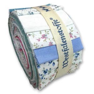 Könnte beinhalten: Ein gerolltes Bündel aus Stoffstreifen in verschiedenen Mustern und Farben, darunter Blumenmuster und einfarbiges Blau. Das Bündel wird von einem cremefarbenen Band mit der Aufschrift "Westfalenstoffe" zusammengehalten. Der Stoff scheint Baumwolle zu sein, geeignet für Quilt- oder Bastelprojekte.