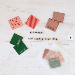 Könnte beinhalten: Eine Sammlung kleiner, quadratischer Geschenkanhänger in verschiedenen Farben, darunter Rosa, Rot und Grün, mit festlichen Designs und Text. Die Anhänger liegen auf einer weißen Holzoberfläche, mit den Worten "FROHE WEIHNACHTEN" in bunten Perlen.