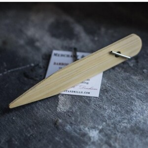 Könnte beinhalten: Ein hellfarbener Pflanzenmarker aus Bambus mit spitzem Ende und Metallstift. Der Marker hat ein Etikett mit dem Text "Merchant & Mills". Der Hintergrund ist eine dunkle, strukturierte Oberfläche.