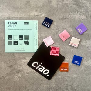 Könnte beinhalten: Eine Sammlung farbiger Stoffetiketten und eine schwarze Karte mit dem weißen Wort "ciao". Die Etiketten zeigen verschiedene Phrasen und Designs, darunter "LOVE", "SCISSORS" und "FASHION PASSION".