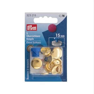 Könnte beinhalten: Eine Packung mit 6 goldfarbenen Überziehknöpfen mit einem Durchmesser von 15 mm. Die Packung enthält ein blaues Plastikwerkzeug zum Anbringen der Knöpfe. Die Packung ist mit "Prym" und "Überziehbare Knöpfe" beschriftet.