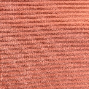 May include: Close-up of a textured, corduroy fabric in a warm, muted terracotta color. The fabric features evenly spaced, parallel ridges running horizontally across the frame, creating a tactile surface.