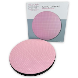 Könnte beinhalten: Eine rosa rotierende Schneidematte mit einem Gittermuster. Die Matte befindet sich auf einer schwarzen Basis und ist für Bastler unterwegs konzipiert. Die Verpackung der Matte befindet sich im Hintergrund und trägt die Aufschrift "Rotating Cutting Mat for Crafters on the Go" und "Sue Daley Designs".