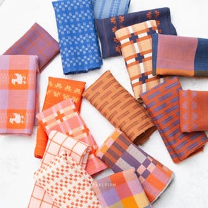 Könnte beinhalten: Eine Sammlung gefalteter Stoffmuster mit verschiedenen Mustern wie Karos, Schachbrettmustern und Blumenmotiven in Orange-, Blau-, Lila- und Rosatönen. Die Stoffstücke sind verstreut angeordnet, wobei der Text FABLEISM deutlich zu sehen ist.