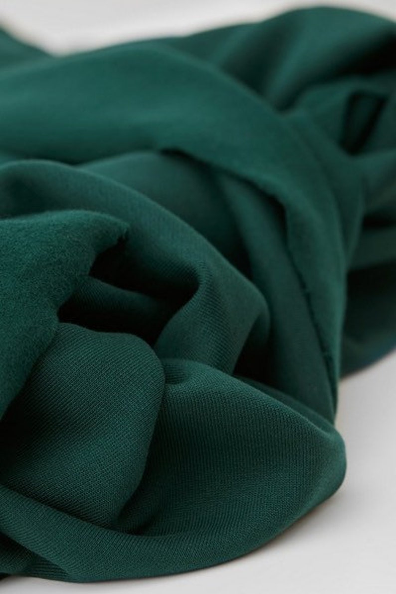 Puede incluir: Primer plano de una tela verde oscuro con una superficie suave y texturizada. La tela est&aacute; doblada y drapeada, mostrando su ca&iacute;da y textura.