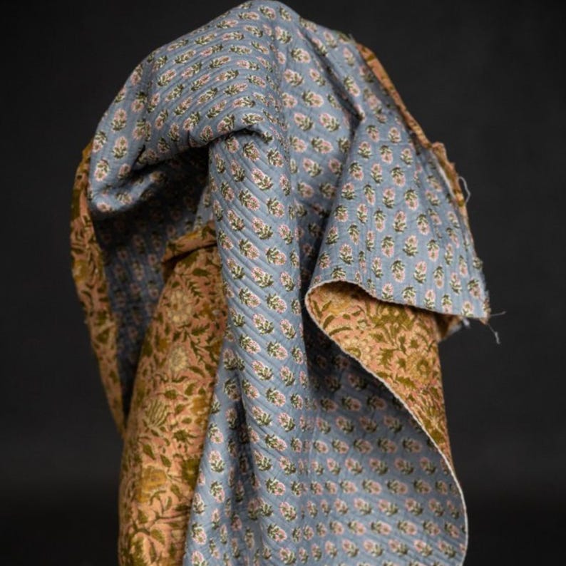 May include: A folded textile featuring a floral pattern in shades of blue, pink, and green. The fabric has a quilted texture and appears to be made of cotton. The design is repeated across the surface, creating a cohesive and visually appealing pattern.