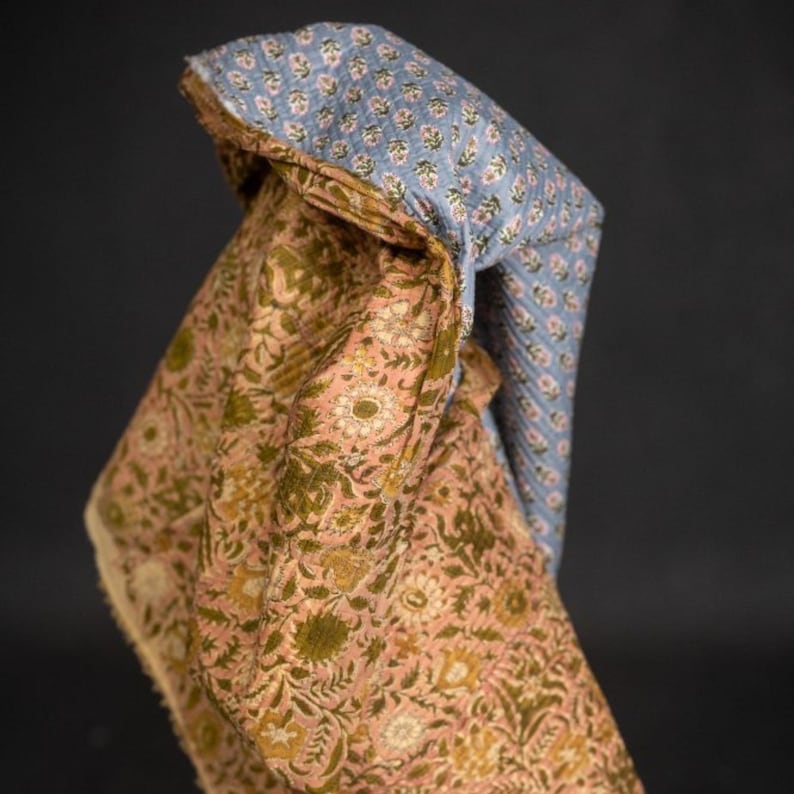 May include: A folded textile featuring two distinct floral patterns. One side displays a light blue fabric with small white flowers, while the other showcases a peach-colored fabric with olive green floral designs. The edges are visible, revealing the layered construction.