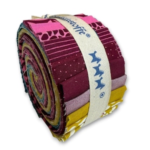 Könnte beinhalten: Ein gerolltes Bündel bunter Stoffstreifen, gehalten von einem cremefarbenen Band. Die Stoffe zeigen verschiedene Muster und Farbtöne von Rosa, Burgunderrot, Gelb und Türkis. Das Band ist mit dem Wort "Moda" und blauen sternartigen Formen bedruckt, was auf einen Bastelbedarf hindeutet.