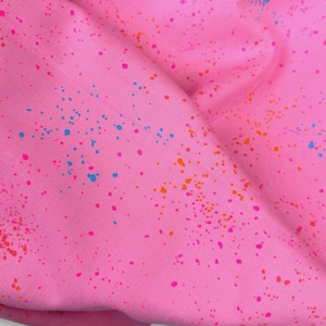 Könnte beinhalten: Nahaufnahme eines rosa Stoffes mit einem gesprenkelten Muster. Der Stoff ist mit kleinen, unregelmäßigen Farbspritzern in Orange, Blau und Rosa bedeckt. Der Stoff ist gefaltet und erzeugt Schatten und Textur.