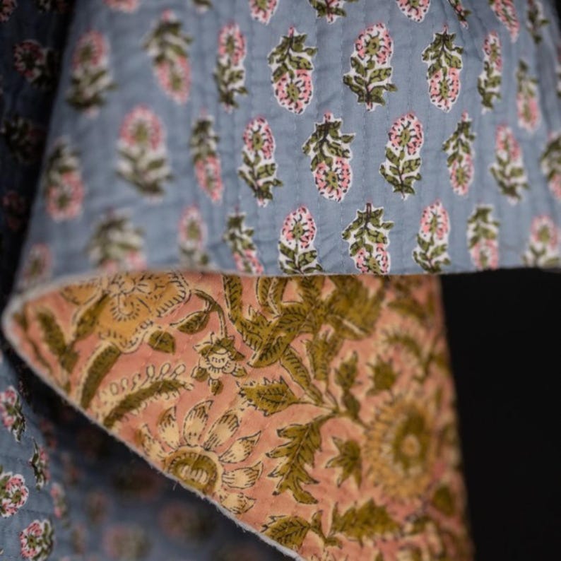 May include: Close-up of a quilted textile with a floral pattern. The top layer is a light blue with pink and green floral designs. The underside is a peach color with large olive green floral patterns. The edges are visible.