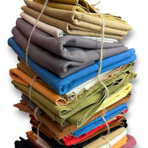 Könnte beinhalten: Ein hoher, vertikaler Stapel gefalteter Stoffstücke in verschiedenen Unifarben, darunter Beige, Grau, Blau, Olivgrün, Gelb, Orange, Rot, Schwarz und Creme. Der gesamte Stapel ist mit einer dünnen, hellen Schnur zusammengebunden.