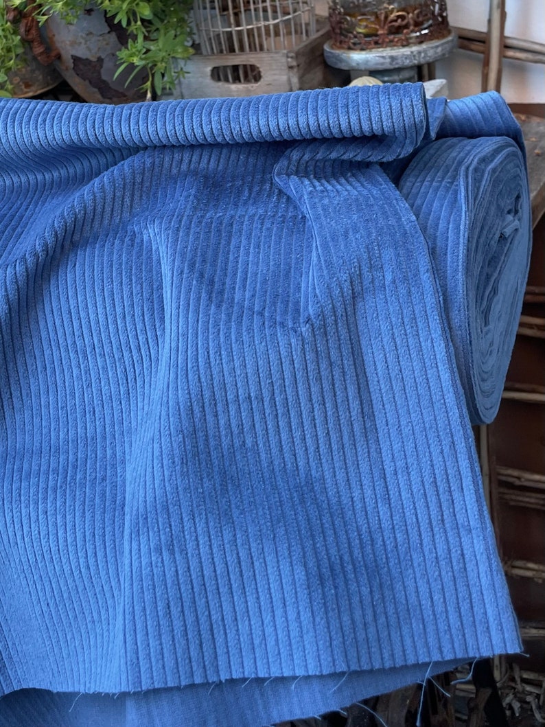 K&ouml;nnte beinhalten: Eine Rolle blauen Cordstoffes mit gerippter Struktur. Der Stoff ist in der Mitte gefaltet und die Rolle ist im Hintergrund sichtbar.