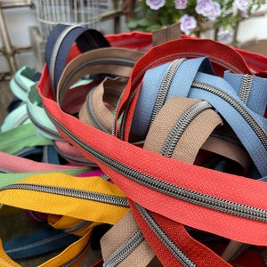 Könnte beinhalten: Eine bunte Auswahl an Reißverschlüssen in verschiedenen Farbtönen, darunter Rot, Blau, Gelb und Grün. Die Reißverschlüsse haben einen metallischen silbernen Zipper und sind in einem Wirrwarr angeordnet, was auf ein Bastel- oder Nähprojekt hindeutet.