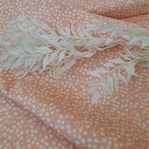 Könnte beinhalten: Pfirsichfarbener Stoff mit einem sich wiederholenden Muster aus kleinen weißen Kreisen und Blättern. Ein weißer, belaubter Zweig liegt auf dem Stoff. Der Stoff wirkt weich und leicht zerknittert.