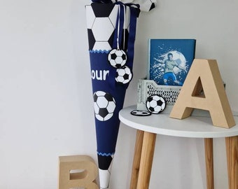 Peut inclure: Un cône scolaire bleu et blanc avec un motif de ballon de football. Le cône est décoré d'un ruban bleu et de breloques en forme de ballon de football. Le cône est posé sur un tapis blanc moelleux avec des lettres dorées qui épellent "12". Il y a aussi des lettres dorées qui épellent "A" et "B" sur la table.