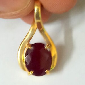 Puede incluir: Un colgante de oro con una gran gema roja oscura de corte ovalado. La gema está sujeta por cuatro garras. El colgante tiene forma de lágrima con un acabado liso y pulido.