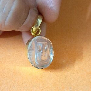 Puede incluir: Un colgante ovalado transparente con un diseño de rostro tallado, engastado en un marco de metal dorado. El colgante tiene una fianza dorada. El colgante está sostenido por una mano sobre un fondo naranja.