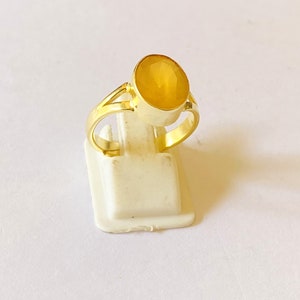 May include: A gold ring with a large, oval-cut yellow gemstone. The ring has a split shank design and is displayed on a white stand against a white background. The gemstone is the focal point, showcasing its colour and cut.