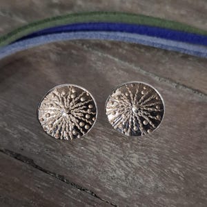 Könnte beinhalten: Ein Paar silberne Ohrringe mit einem runden Design, das ein strukturiertes, strahlendes Muster aufweist. Die Ohrringe haben eine leicht erhöhte Mitte und eine glatte, polierte Oberfläche.