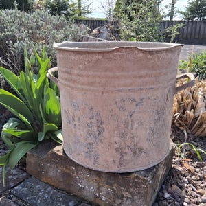 May include: A weathered, cylindrical metal bucket with two handles. The bucket has a rustic, aged appearance with visible rust and wear. It sits on a brick base, surrounded by greenery and plants, suggesting outdoor use for gardening or storage.