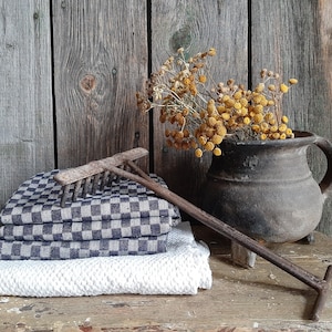 May include: A rustic, dark brown clay pot with a handle sits on a wooden surface. The pot is filled with dried yellow flowers. A vintage-style rake with a wooden handle lies on a stack of blue and white checked fabric and a white towel.