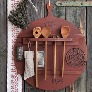 May include: A rustic, circular, red-painted wooden kitchen organizer with a handle. It features wooden spoons, graters, and a cooling rack. The organizer is inscribed with "A.B." and "1895". A decorative floral ribbon and dried herbs hang on the left.