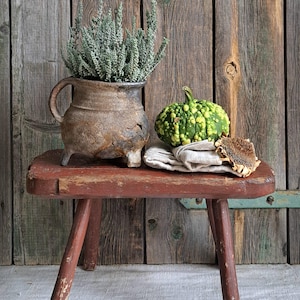 Puede incluir: Naturaleza muerta rústica con un pequeño taburete de madera rojo desgastado. Una maceta de cerámica marrón texturizada con asa contiene una planta verde. Una calabaza verde con bultos y una flor seca descansan sobre un paño de lino.