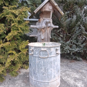 Könnte beinhalten: Ein rustikales Vogelhaus mit Holzdach und einem verwitterten Baumstammfuß, montiert über einem großen, zylindrischen Metallbehälter. Der Behälter hat Griffe und eine abgenutzte, graue Oberfläche, vor dem Hintergrund von immergrünem Laub.