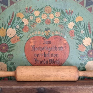 Può includere: Un mattarello di legno con manici poggia su una superficie di legno. Lo sfondo presenta un pannello di legno dipinto e antico con motivi floreali e a forma di cuore nei toni del verde, arancione e rosso. Il testo è visibile sul cuore.