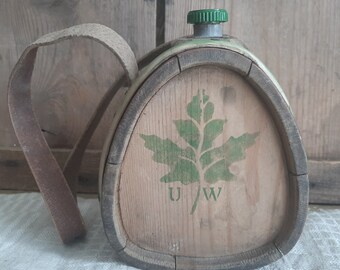 May include: A wooden canteen with a green leaf design and the letters "U/W" printed on the front. The canteen has a brown leather strap and a green metal lid.