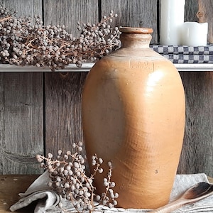 May include: A large, antique, tan-colored stoneware jug with a narrow neck and a rounded body. The jug is displayed on a wooden surface with a wooden spoon, dried berries, and a folded linen cloth. Two white candles sit on a shelf in the background.