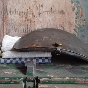 May include: A vintage metal canteen with a hinged lid and a brass cap. The canteen is resting on a wooden surface with a blue and white checked cloth.
