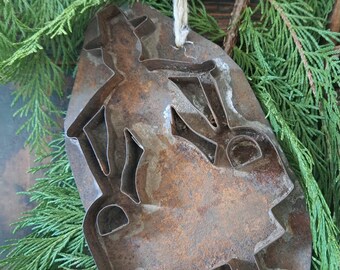 Op de afbeelding: Een roestige metalen koekjesvorm in de vorm van een cowboy, met een touwtje om op te hangen. De vorm heeft een verweerde, bruine afwerking en is geplaatst tegen een achtergrond van groene bladeren.
